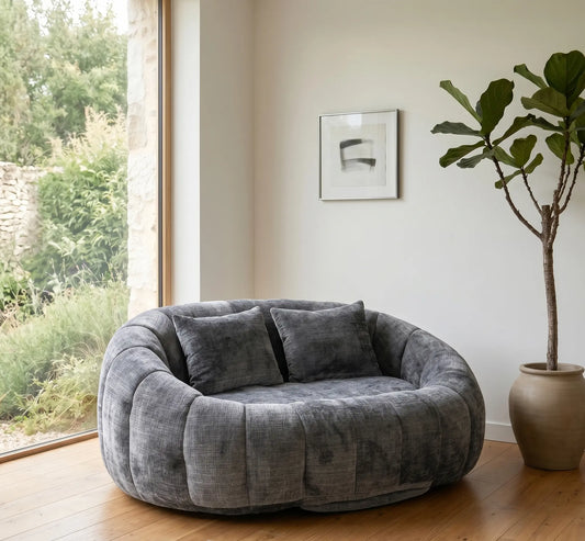 Ambiance salon zen avec un grand canapé pouf panoramique gris anthracite, parquet en chêne, plante verte Ficus et grande baie vitrée ouvrant sur un jardin.