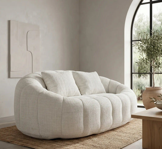 Aménagement d'un salon minimaliste et lumineux avec un grand canapé pouf panoramique blanc crème, un tapis texturé assorti et un décor épuré avec pampas.